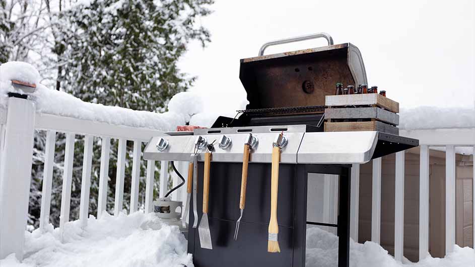 Un barbecue gris avec des outils est sur une terrasse enneigée, avec des arbres en arrière-plan.