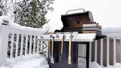 Un barbecue gris avec des outils est sur une terrasse enneigée, avec des arbres en arrière-plan.