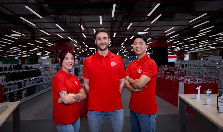 Trois employés en polos rouges posent en souriant dans un magasin d’électronique, avec rayons et plafonniers lumineux derrière eux.
