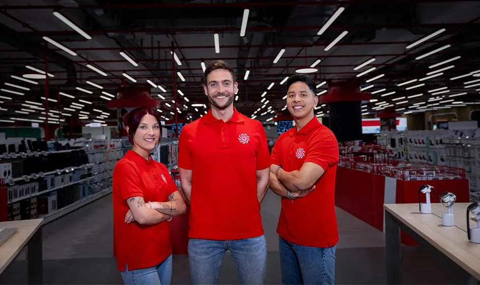 Trois employés en polos rouges posent en souriant dans un magasin d’électronique, avec rayons et plafonniers lumineux derrière eux.