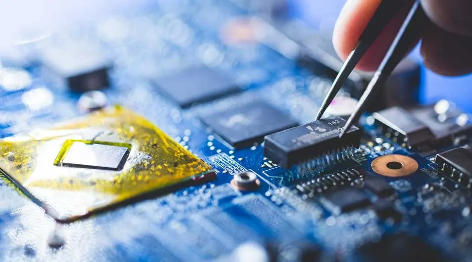Detailopname van een blauwe printplaat met elektronische componenten, waaronder een goudkleurige chip, en een pincet dat een kleine zwarte chip vasthoudt.
