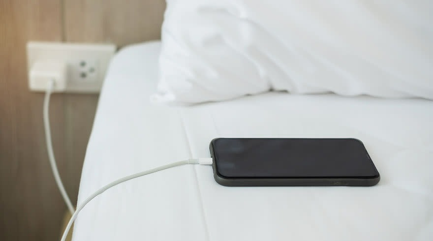 Téléphone noir en charge sur un lit blanc près d'une prise murale.