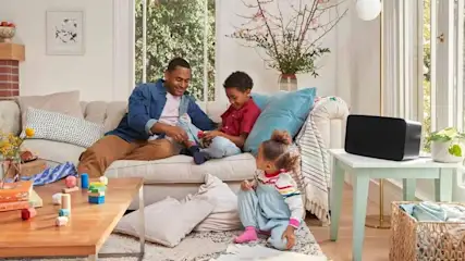Un salon lumineux avec un canapé beige, une table en bois, et une enceinte noire sur une table d'appoint vert clair.