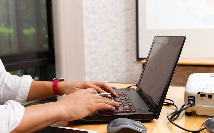 Une personne tape sur un ordinateur portable relié par câbles à un projecteur sur un bureau, prête pour présenter.