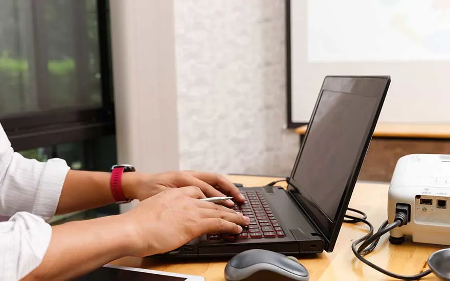 Une personne tape sur un ordinateur portable relié par câbles à un projecteur sur un bureau, prête pour présenter.