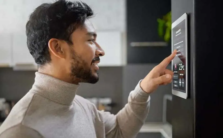 Un écran tactile affiche 23°C sur un réfrigérateur noir. Une main interagit avec l'écran. Le fond est une cuisine.