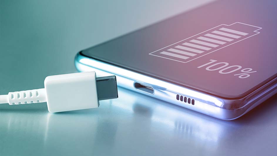 Een zilveren smartphone ligt op een blauwe ondergrond, met een witte USB-C kabel aangesloten. Het scherm toont een batterij-icoon en "100%".