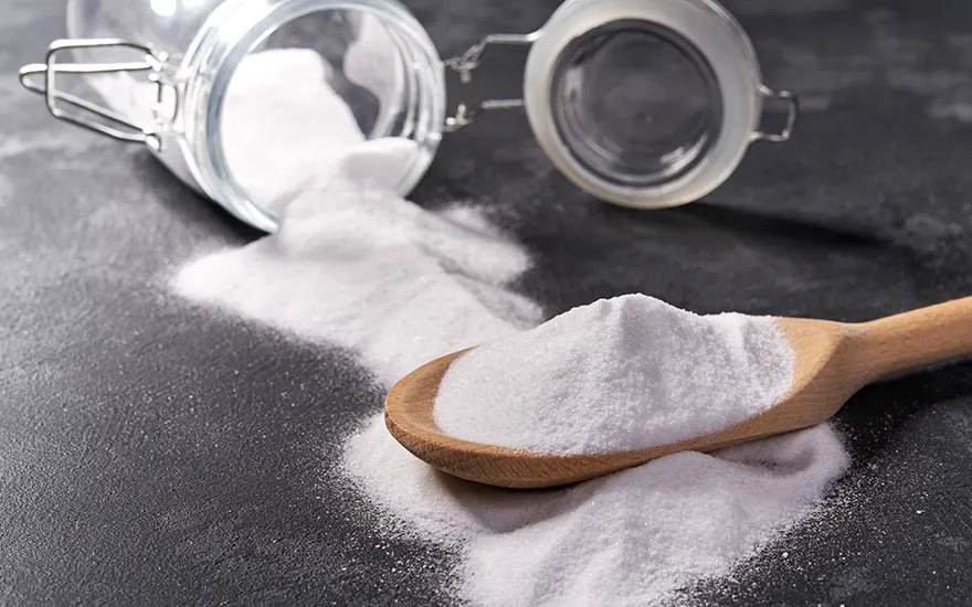 Un bocal en verre ouvert est renversé, déversant une poudre blanche sur une cuillère en bois, sur une surface sombre.