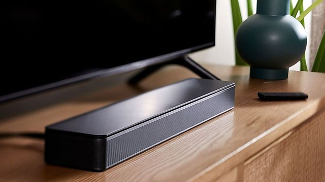 Une barre de son noire est placée devant un téléviseur sur une table en bois, avec un vase vert et une télécommande à côté.