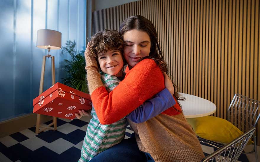 Een vrouw omhelst een glimlachende jongen die een rood cadeau vasthoudt in een warme, gezellige woonkamer.