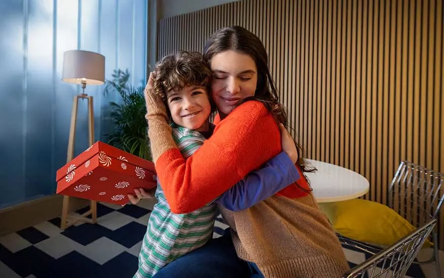 Een vrouw omhelst een glimlachende jongen die een rood cadeau vasthoudt in een warme, gezellige woonkamer.