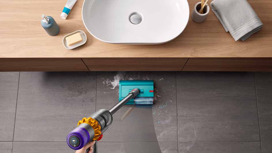 Vue plongeante sur un sol carrelé gris nettoyé avec un aspirateur bleu et violet. Un lavabo et des articles de toilette sont au-dessus.