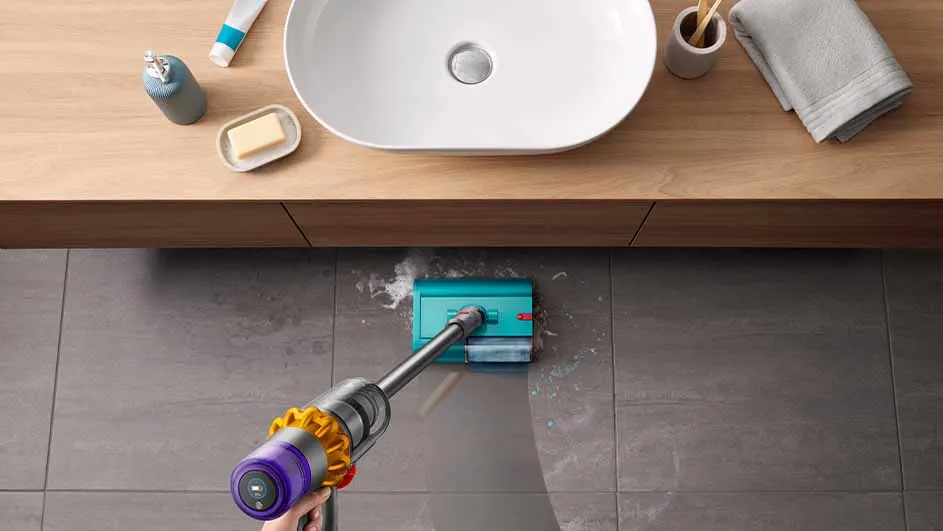 Vue plongeante sur un sol carrelé gris nettoyé avec un aspirateur bleu et violet. Un lavabo et des articles de toilette sont au-dessus.