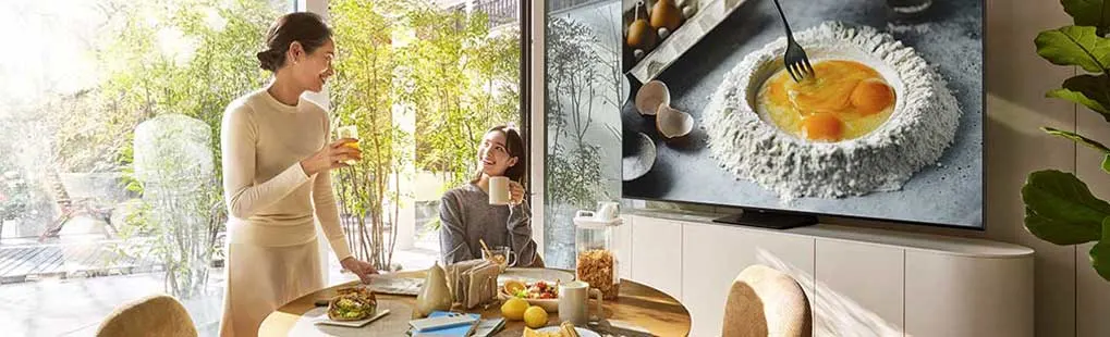 Twee mensen ontbijten aan een ronde tafel met fruit, brood en sap. Op de Samsung-tv staat een kookprogramma met eieren en bloem.