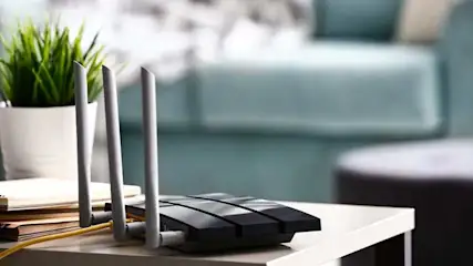 Een zwarte router met drie witte antennes op een wit tafeltje, naast een plant en boeken. Een blauwe bank is op de achtergrond te zien.