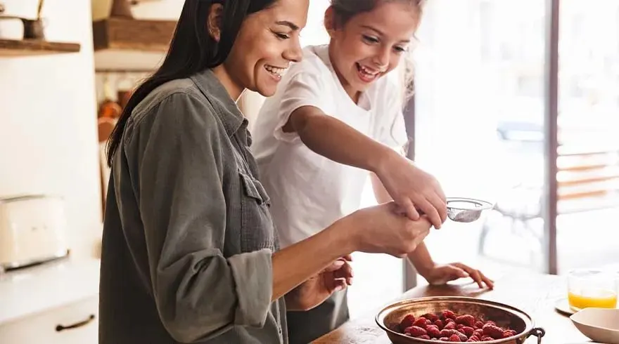 Une femme et une fille préparent des framboises dans une cuisine lumineuse avec des appareils blancs et des étagères en bois.