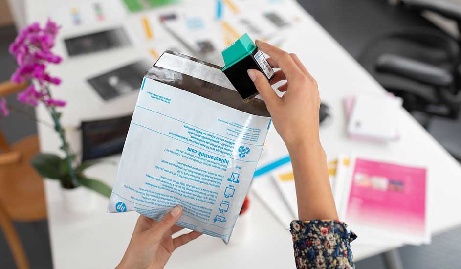 Iemand haalt een groene cartridge uit een witte HP-envelop op een witte tafel met bloemen.