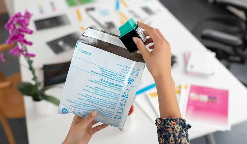 Iemand haalt een groene cartridge uit een witte HP-envelop op een witte tafel met bloemen.