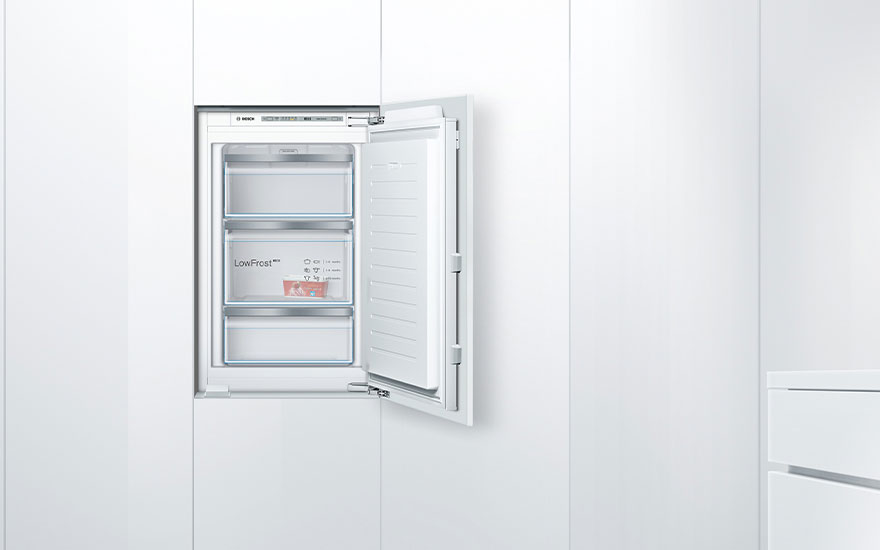 Congélateur blanc intégré, porte ouverte, avec trois tiroirs partiellement remplis, dans une armoire de cuisine blanche.