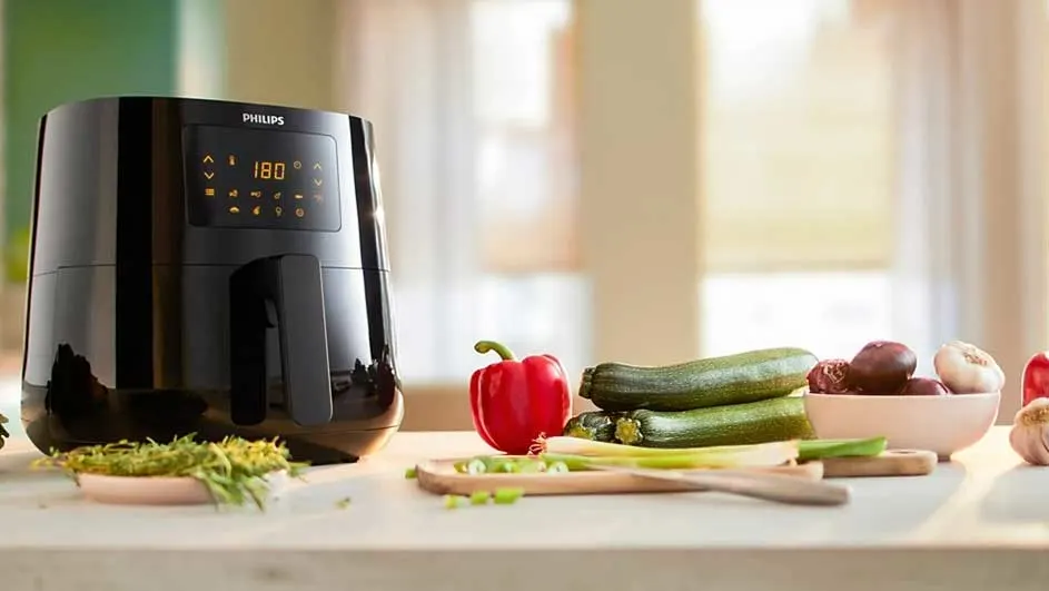 Une friteuse Philips noire à écran 180°, des légumes(courgettes, poivron rouge, oignons) et de l'ail sur une table de cuisine.
