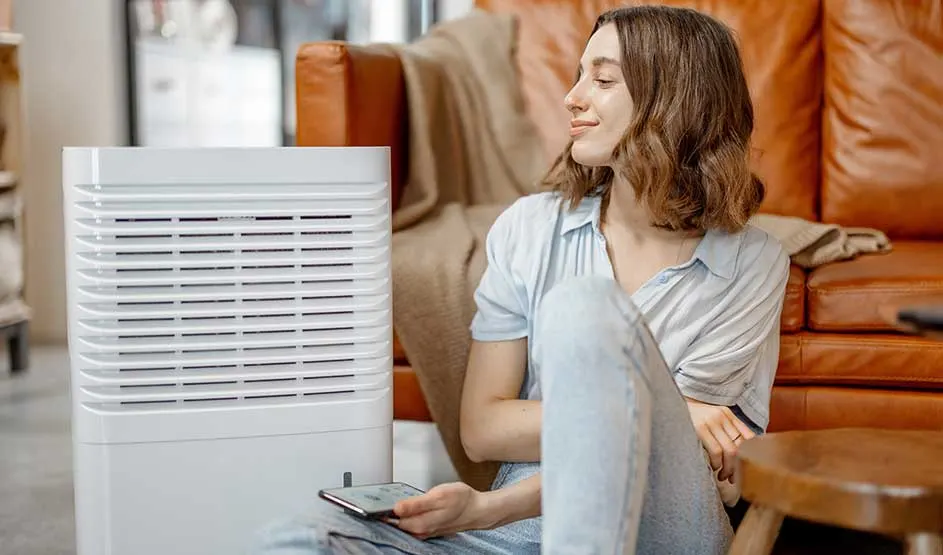 Vrouw zit relaxed op de vloer bij een bruine leren sofa, glimlachend naar een witte luchtreiniger naast haar, smartphone losjes in de hand.