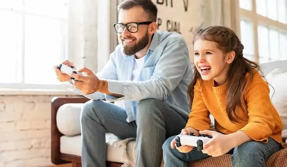 A smiling father wearing glasses and his daughter in an orange jumper play video games together with controllers in a bright modern lounge.