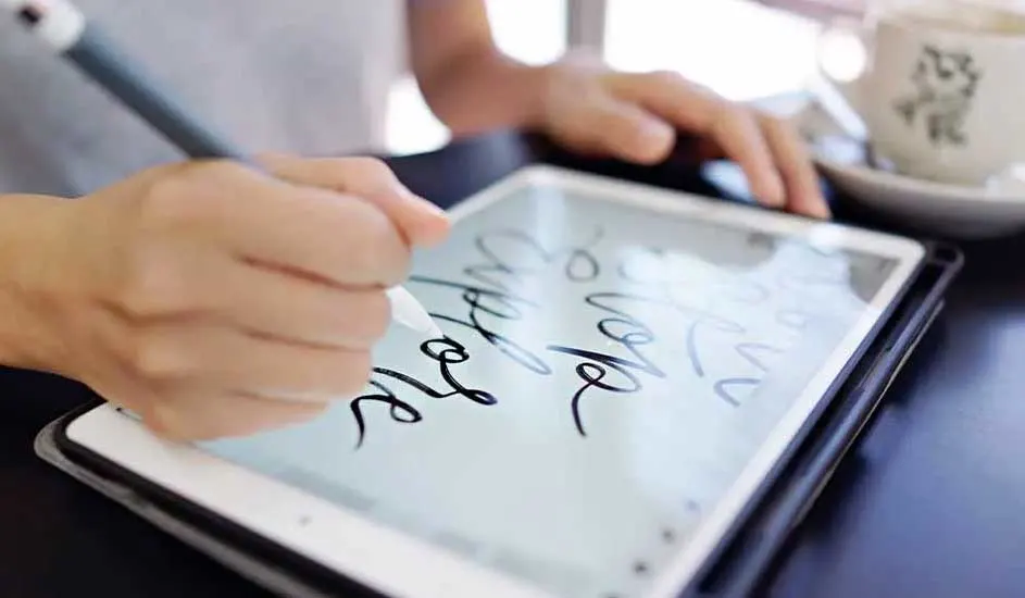 Hands use a stylus on a tablet displaying script. A white mug sits on a saucer in the background.