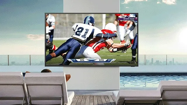 Een tv toont een American Football wedstrijd naast een zwembad met witte stoelen. De lucht is blauw.