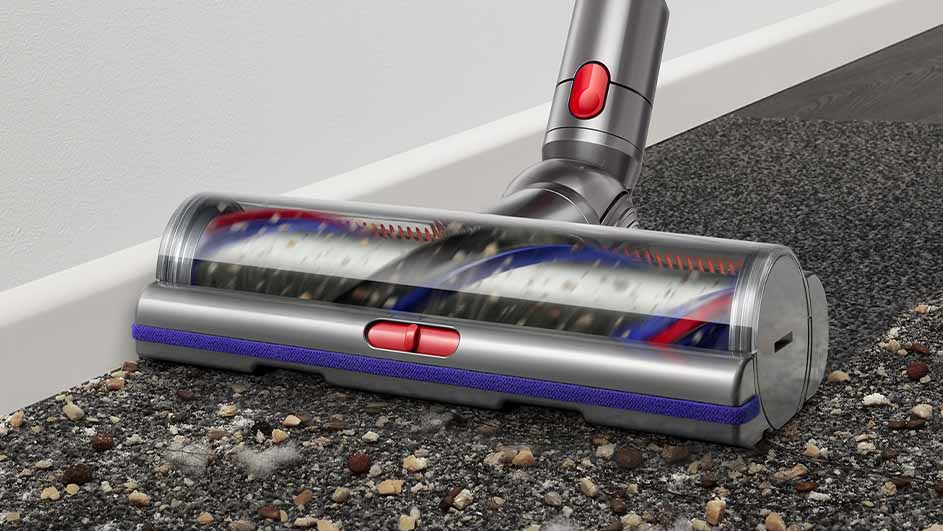 Aspirateur Dyson argenté sur un tapis texturé gris avec des débris, près d'une plinthe blanche. Détails rouges et bleus visibles.