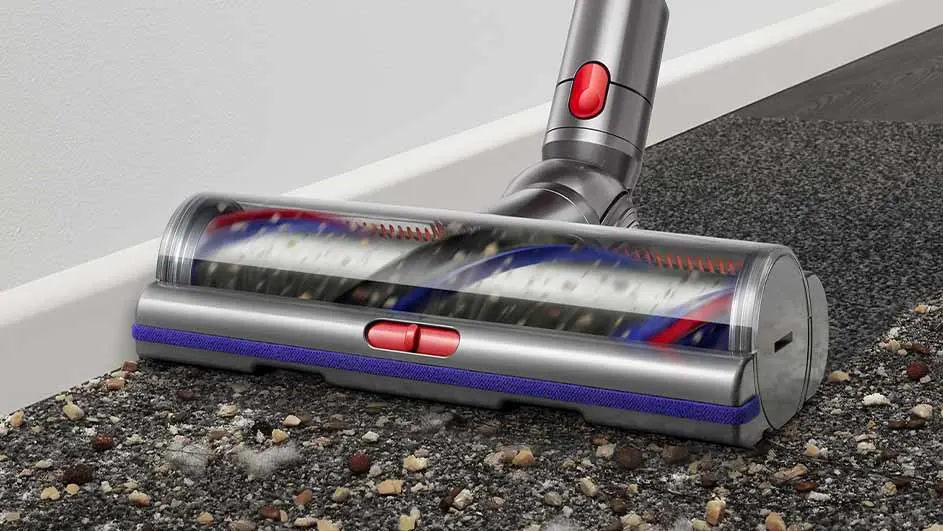 Aspirateur Dyson argenté sur un tapis texturé gris avec des débris, près d'une plinthe blanche. Détails rouges et bleus visibles.