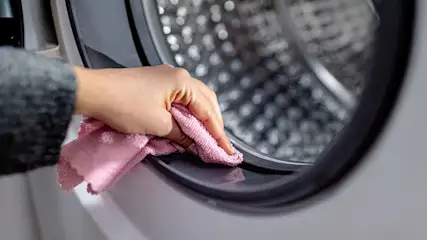 Een roze doek reinigt de grijze rubberen afdichting van een witte wasmachine, met een metalen trommel zichtbaar.