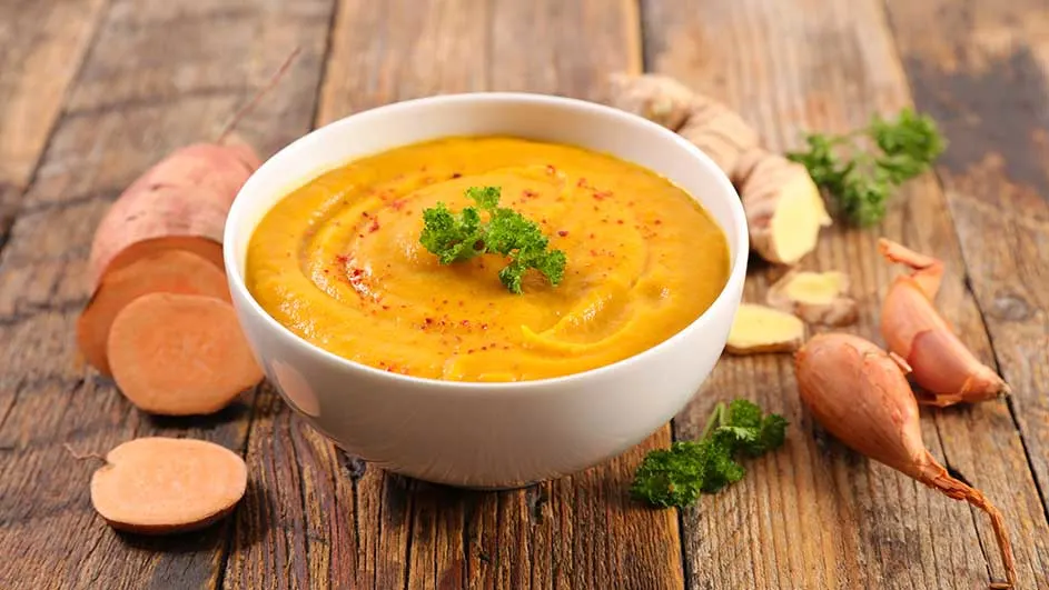 Bol de soupe orange sur bois, garni de persil. Patate douce, gingembre et échalotes autour.