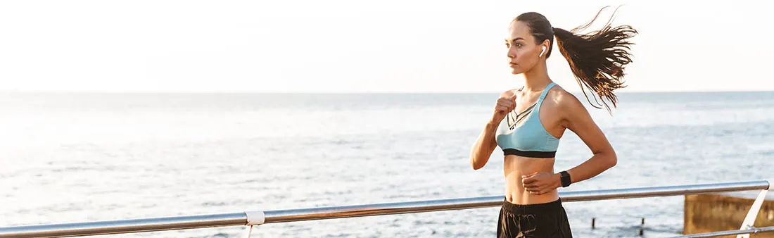 Une femme fait du jogging en bord de mer avec un soutien-gorge de sport bleu et un short noir. Une rampe métallique est visible.