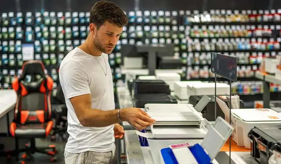 Een man staat bij een rij printers in een winkel. Er is een rode bureaustoel en rekken vol producten op de achtergrond.