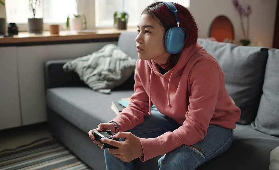 A person with blue headphones sits on a grey couch, holding a black game controller, wearing a pink hoodie and jeans. Windows are in the background.