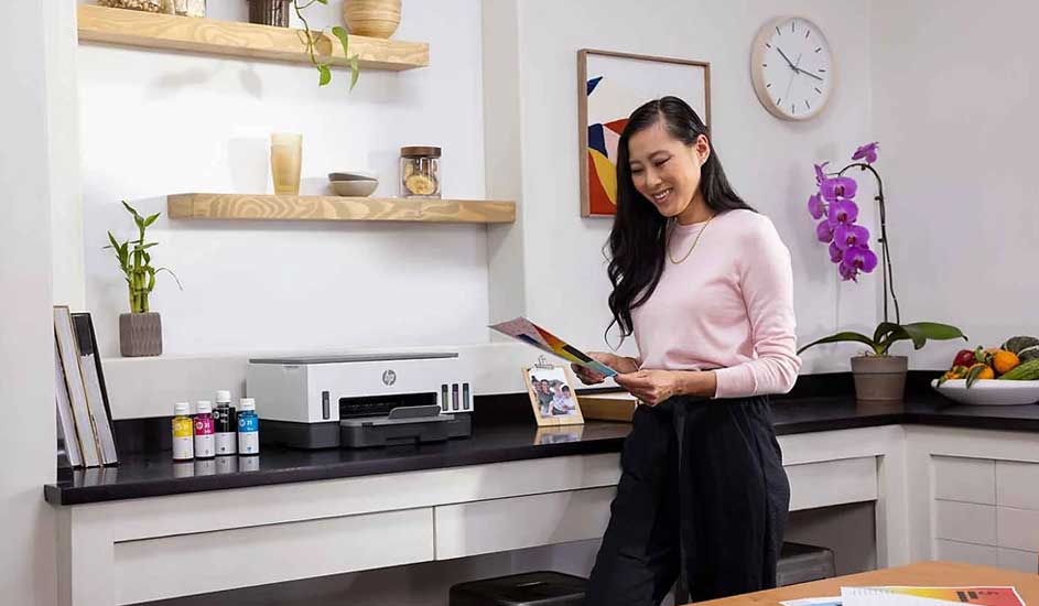 A printer sits on a black counter next to ink bottles. Wooden shelves are on the wall. An orchid and bowl of fruit sit on the counter.