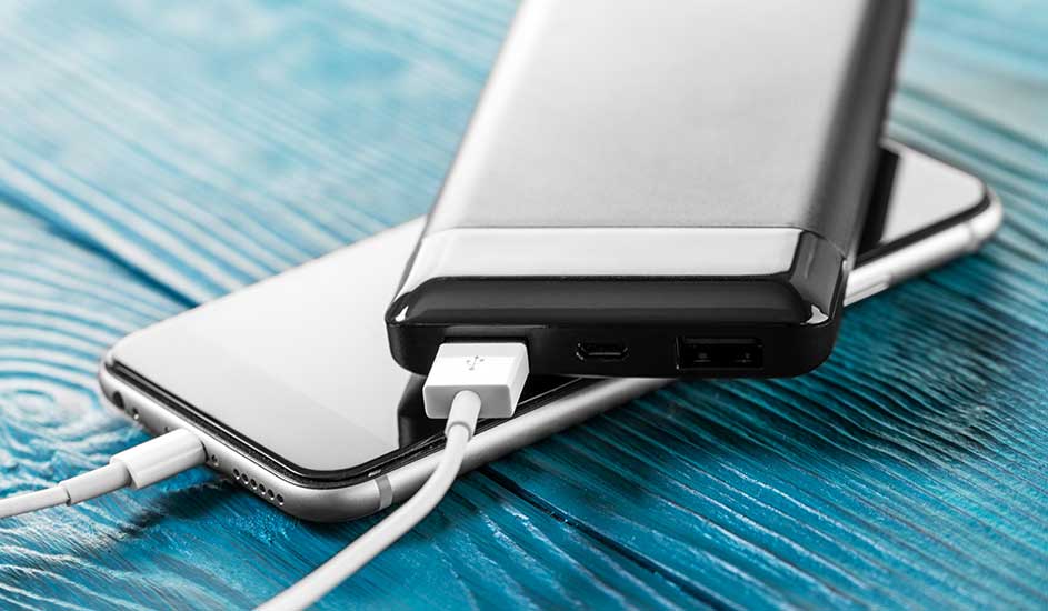 Een zilverkleurige telefoon wordt opgeladen door een grijze powerbank met een witte kabel op een blauwe houten ondergrond.