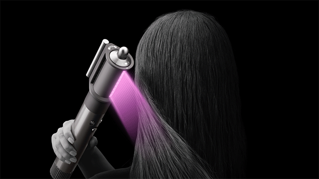 Een hand houdt een stylingtool tegen donker haar, met een paarse lichtstraal die op het haar schijnt, tegen een zwarte achtergrond.