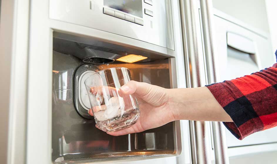 Une main tient un verre sous le distributeur d’un réfrigérateur moderne, qui verse de la glace ou de l’eau fraîche dans le récipient.