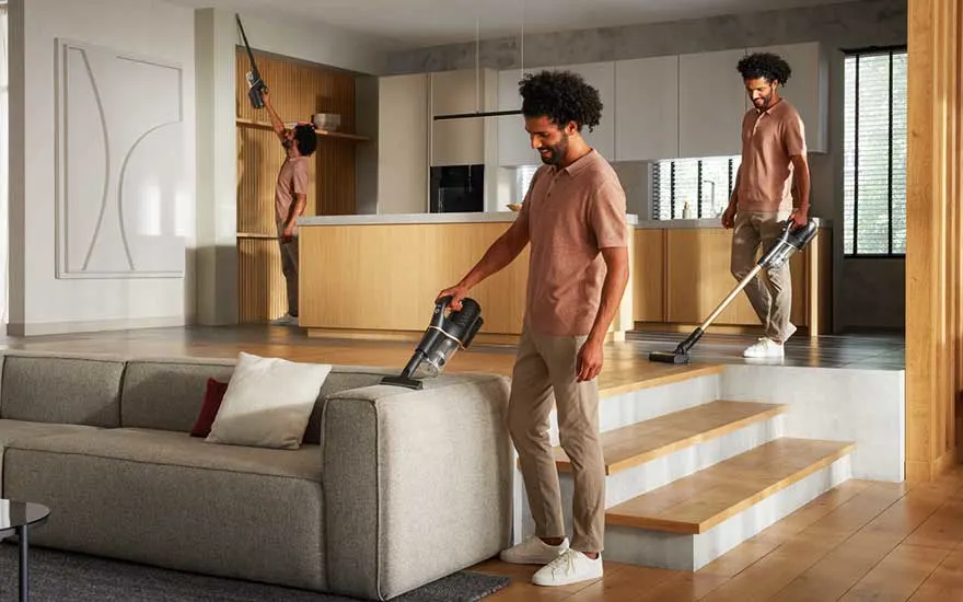 Een moderne woonkamer met een grijze bank en houten vloeren. Een stofzuiger wordt gebruikt.