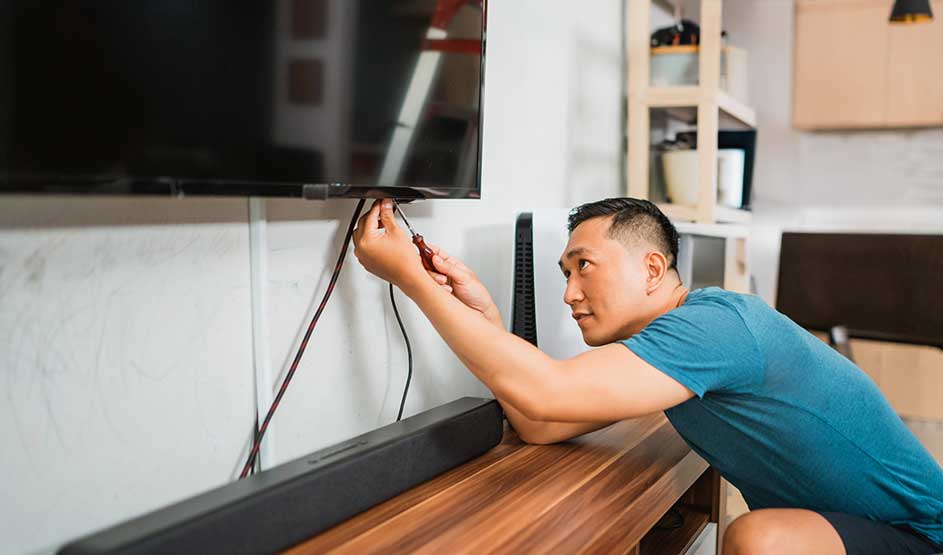 A man connects cables beneath a wall-mounted television in a modern living room with a soundbar.