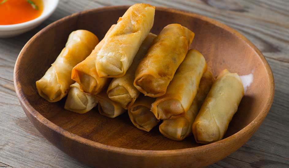 Assiette en bois remplie de rouleaux de printemps dorés. Un bol de sauce orange est visible à l'arrière-plan.