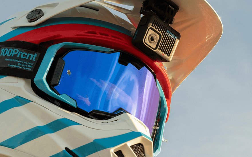 Close-up of a white helmet with blue stripes, red trim, and blue goggles. A camera is mounted on top. The background is a light blue sky.
