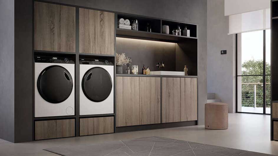 Een wasruimte met twee wasmachines naast een houten kast met een aanrecht en open schappen. Een raam laat daglicht binnen.