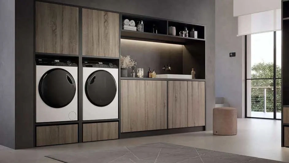 Een wasruimte met twee wasmachines naast een houten kast met een aanrecht en open schappen. Een raam laat daglicht binnen.