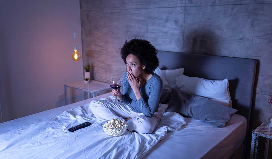 Vrouw zit rechtop in bed, kijkt gespannen naar tv, met kom popcorn, glas wijn en afstandsbediening naast zich.