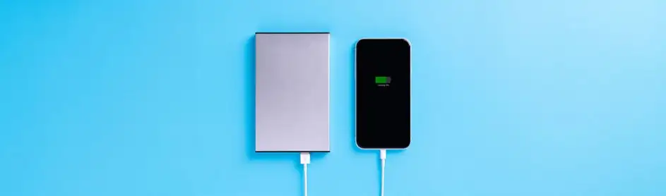 Un chargeur portable gris est connecté par un câble blanc à un smartphone noir sur un fond bleu clair. L'écran affiche une batterie verte.