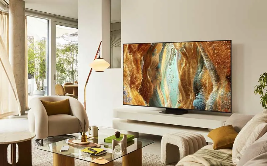 Een lichte woonkamer met grote tv op een laag meubel, beige bank, fauteuil, glazen salontafel, planten en ramen met zachte gordijnen.