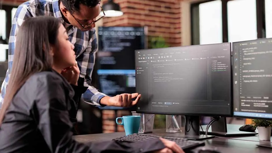 Op een bureau staan twee computerschermen met code, een toetsenbord, een blauwe mok en een kleine plant. Bakstenen muur en raam op achtergrond.