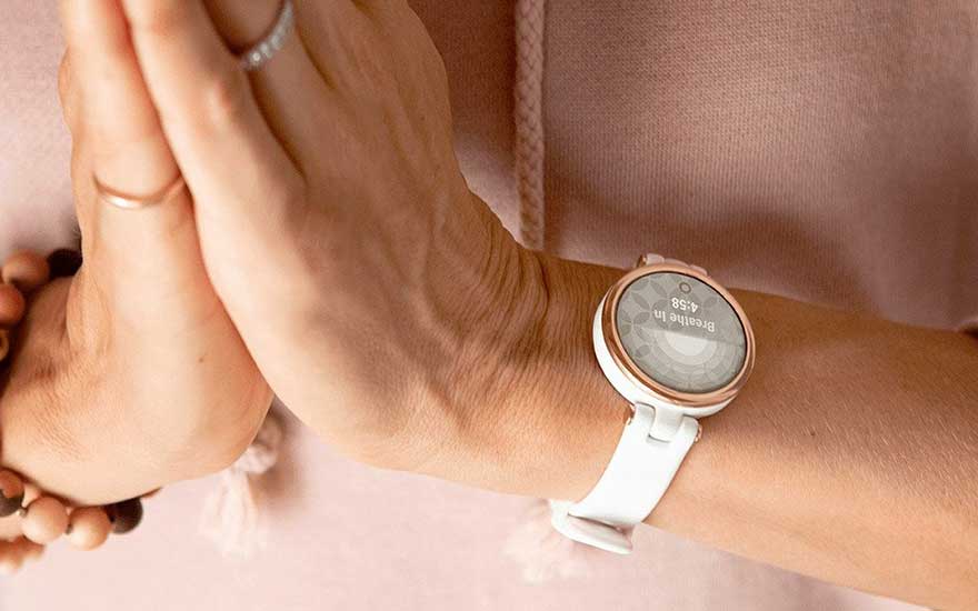 Une montre connectée blanche et or affichant "Breathe In 4:58" est portée sur le poignet, avec des mains jointes et un bracelet en perles.